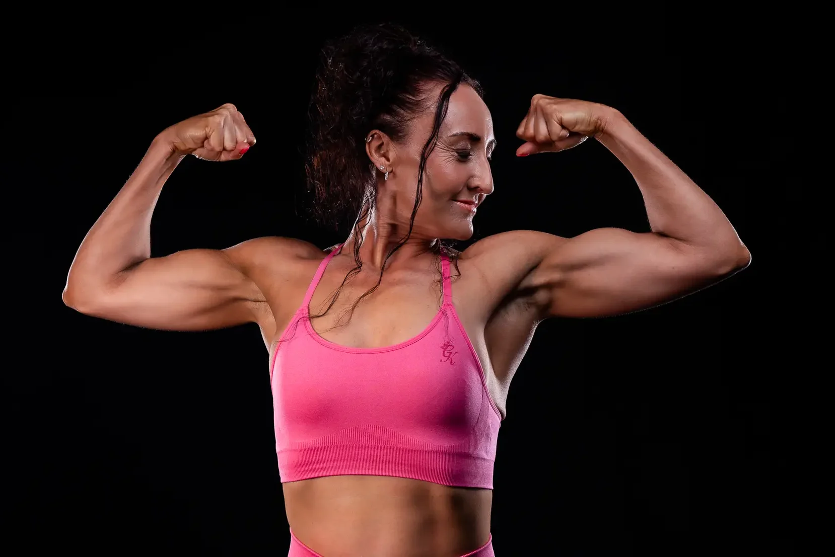 A fit woman in a pink sports bra confidently flexing her biceps, standing against a black background